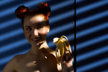 Creative portrait of a red-haired girl with a banana in her hands on a blue background with a stripe from the sun's shadow - 355212919