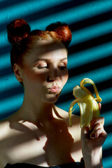 Creative portrait of a red-haired girl with a banana in her hands on a blue background with a stripe from the sun's shadow - 355213138