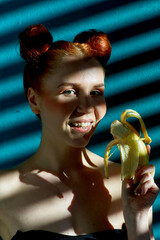 Creative portrait of a red-haired girl with a banana in her hands on a blue background with a stripe from the sun's shadow - 355213164