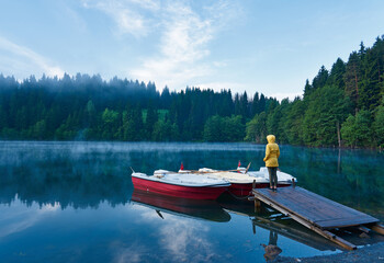 A girl in yellow coat is standing and posing on the wooden dock near boats on the foggy lake...