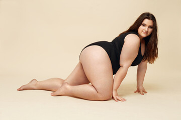 Obraz na płótnie Canvas Plus Size Model. Full-Figured Woman In Black Bodysuit Portrait. Crawling Brunette Posing On Beige Background And Looking At Camera. Body Positive Concept.