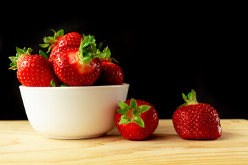 White bowl with fresh strawberries on a wooden Board on a black background. Next to the bowl are two strawberries