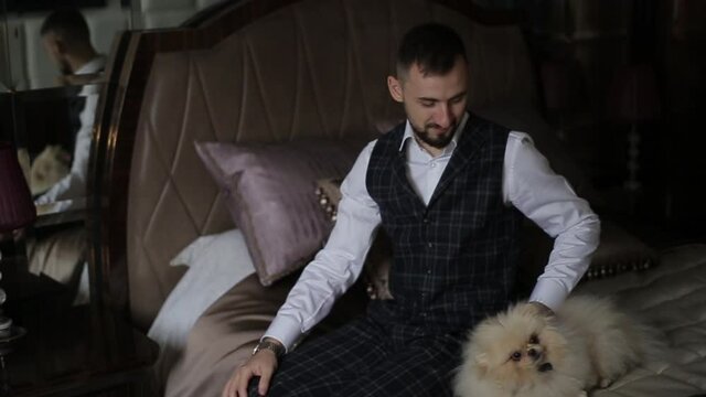 Handsome young bearded man in a stylish suit sits on a luxurious bed in a hotel in the morning and caresses his beloved dog. The kind groom smiles and strokes the spitz on the bed in the bedroom.