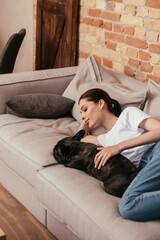 smiling woman lying on sofa and touching black french bulldog