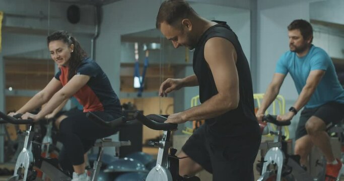 Handsome Caucasian sportsman joining group of athletic people on cycling equipment. Positive young man greeting men and women in gym riding on exercise bikes. Cinema 4k ProRes HQ.