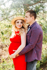 Fototapeta premium Pregnant girl with a tummy, awaiting delivery, in a red dress and a straw hat with her husband in a field and feather grass