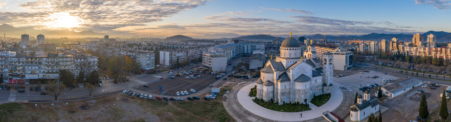 Fototapeta na wymiar Podgorica, Montenegro: Cathedral of the Resurrection of Christ at sunrise. Aerial shot of the famous landmark including panorama of the city in the first morning light under orange and blue sky.