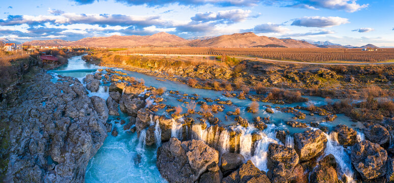 Cascades and whitewater rapids with frothy water at the waterfall Niagara on Cijevna (Cem) river near Podgorica and Tuzi in Montenegro. Large vineyard and Decic hill, at sunset, under beautiful sky.