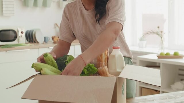 Medium shot of young mixed-race woman standing at kitchen table, taking fresh vegetables out of delivered box and putting them on table