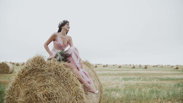 Carefree lady in dress posing for camera on a haystack in windy field