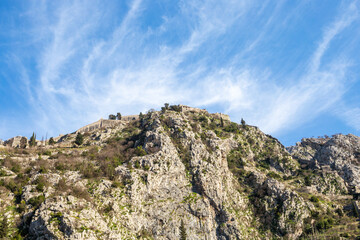 Mountain ridge featuring a medieval fortification wall under blue sky with high cloud formations. Rough and rocky terrain in the Mediterranean. Hills overlooking Kotor Bay in Montenegro.