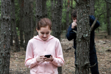 A maniac watches from behind a tree in the forest for a teenage girl in a tracksuit with a mobile phone in her hands. The concept of child abduction.
