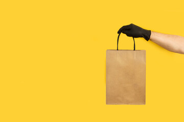 Mano de hombre con guante de látex negro sosteniendo una bolsa de papel sobre un fondo amarillo...