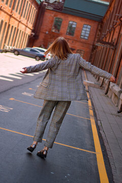 Unrecognizable blond lady wearing plaiding trouser suit going and jumping anong the street with red brick buildings in positive mood. Shot from the back, Faceless trend. Angled horizon. Dinamics enjoy