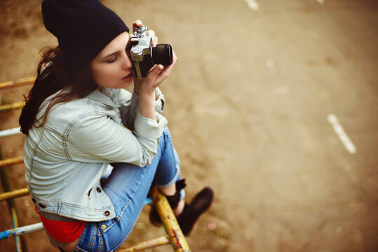 portrait of a beautiful hipster girl with a camera taking photos and sitting on a children's slide in the park outdoor
