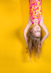 Excited crazy little blonde girl in a swimsuit, hanging upside down on a yellow studio background....