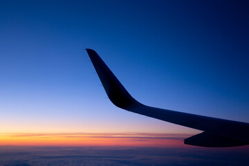 Silhouette of the left wing and winglet of an aircraft at the cruising altitude above the clouds early in the morning, at dawn, with clear blue sky and orange horizon as the backdrop. Red-eye flight.