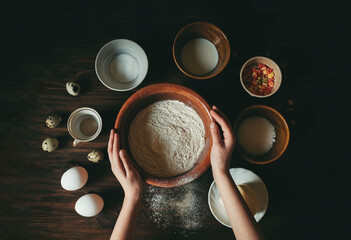 hands holding a clay pot with flour on a dark wooden table and around ingredients for making bread such as eggs butter sugar milk