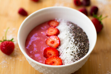 Healthy vegan trendy breakfast: magenta pink berry smoothie bowl made with frozen bananas, raspberry, strawberry and coconut milk. Topped with coconut flakes chia seeds and sliced strawberry. 