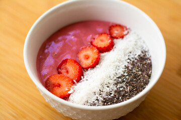 Healthy vegan trendy breakfast: magenta pink berry smoothie bowl made with frozen bananas, raspberry, strawberry and coconut milk. Topped with coconut flakes chia seeds and sliced strawberry. 