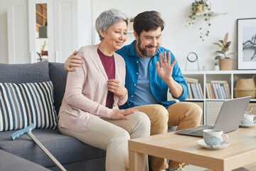 Mature man sitting on sofa with senior woman and waving his hand they communicating online on laptop