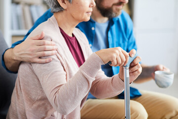 Close-up of senior woman holding crutch and sitting on the sofa with male nurse embracing her and sitting nearby
