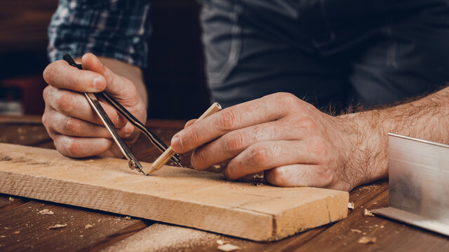 A close up of a male carpenter marks with a metal compass and pencil on a wooden bar.