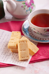 A delicious cup of black tea in an ornate porcelain cup with some delicious confectionary next to the cup and a teapot in the background.