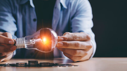 New idea concept with coins stack save power to save the earth, young Asian men hand holding light bulb  with new knowledge light bulb and money stack on wood table on black background with copy space