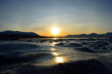Fototapeta na wymiar golden sunrise over blue fjord and snowy mountain with reflection on thick frozen sea shore ice