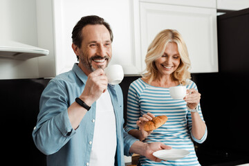 Cheerful middle aged couple drinking coffee and holding croissant in kitchen