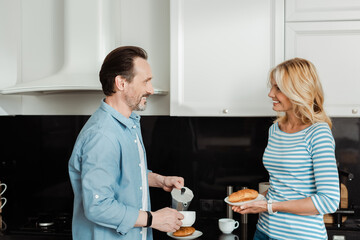 Smiling man pouring coffee near beautiful wife holding croissant in kitchen