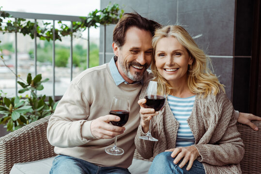 Smiling mature couple holding wine glasses on wicker sofa on terrace
