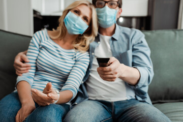 Selective focus of mature couple in medical masks watching tv at home