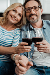 Selective focus of smiling mature couple holding hands and clinking with wine at home