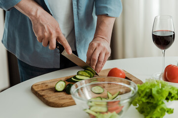 Cropped view of man cutting cucumber while cooking salad near glass of wine in kitchen