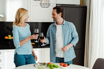 Smiling mature couple clinking with wine near fresh vegetables on kitchen table