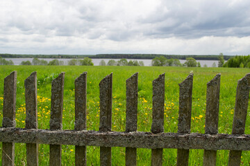 An old wooden fence covered with moss against the background of a meadow, river and sky with clouds.