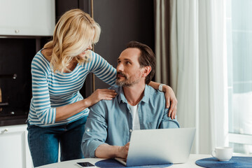 Selective focus of smiling woman embracing handsome husband using laptop on kitchen table