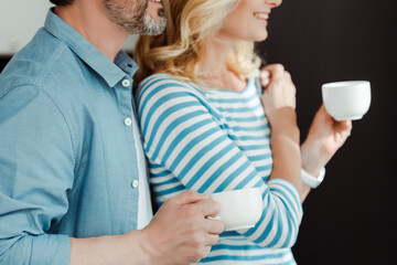 Cropped view of man embracing smiling wife with coffee cup at home