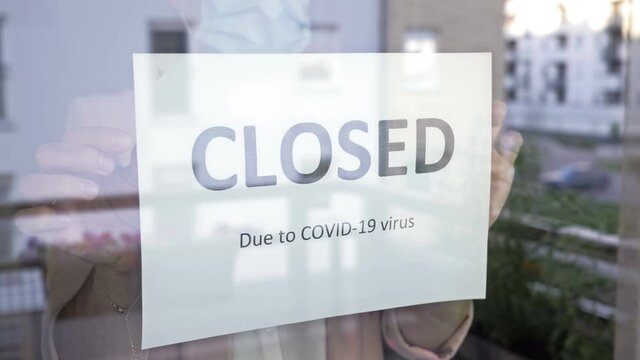 Business office or store shop is closed, bankrupt business due to the Coronavirus pandemic. Unidentified person in a medical mask attach the sign CLOSED DUE TO COVID-19 on the glass door.