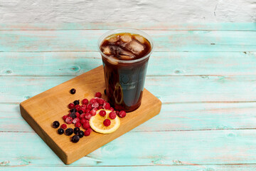 cold coffee summer drink brown with ice lemon and berries. Drink in a clear glass in a still life with red and black berries on a light blue wooden background. beautiful still life with a cold drink