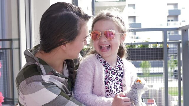 Young woman sits with her six-year-old daughter on the balcony. Mom and chaild are a lot of fun. Photographer is reflected in the mirror glasses of the girl.