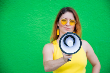 Fototapeta premium Happy woman in a yellow dress glasses and earrings on a green background shouting into a megaphone. Portrait of a girl holding a loudspeaker. Lady with a perfect snow-white smile.