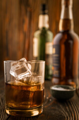 A glass of whiskey with ice, in the background are bottles on a wooden table of a bar counter, shallow depth of field, selective focus. The concept of alcoholic drinks in a roadside bar.