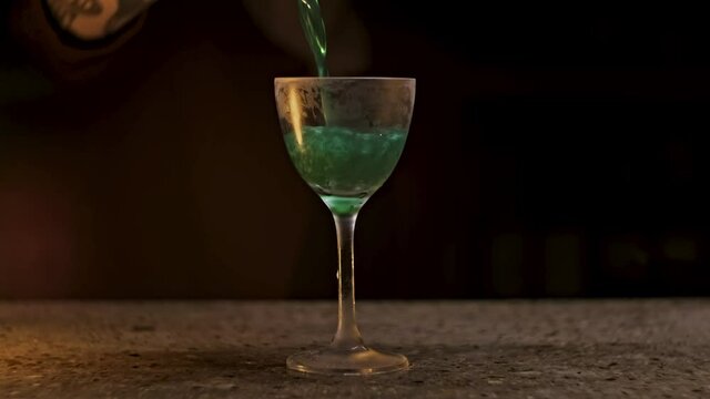 Wine glass with green coctail on black background. Stock footage. Close up of a hand pouring alcoholic beverage of green color with shining particles.