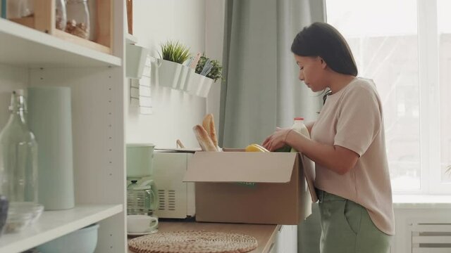 Medium shot of beautiful mixed-race woman carrying box with fresh vegetables and bread, putting it on kitchen worktop and starting taking food out of it