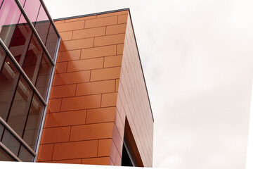 Orange coloured aluminum metal composite panels on the corner of a building with closed glass windows. The background is of white clouds. 