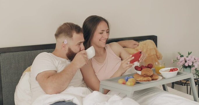 Happy young couple talking and smiling while looking at phone screen. Man drinking coffee while woman holding smartphone during breakfast in bed. Concept of relationship and leisure.