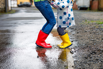 Close-up of two children, toddler girl and kid boy wearing red and yellow rain boots, walking during sleet. Happy siblings, brother and sister jumping into puddle. Having fun outdoors, active family